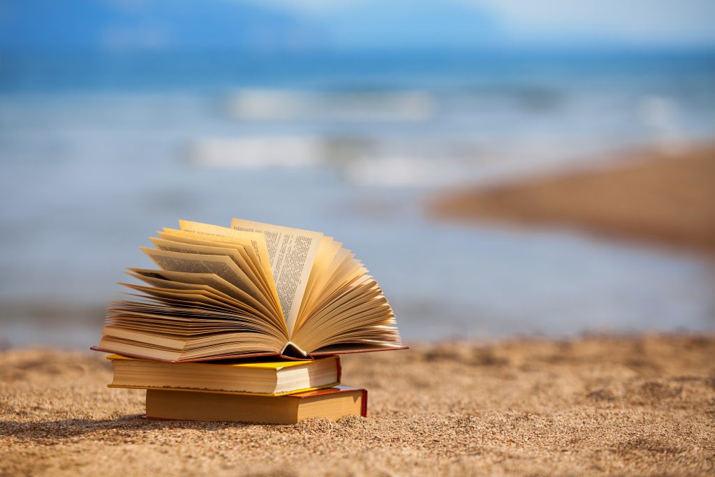 stack of books on the sand with the sea in the background. The top book is open with the pages flapping in the breeze.