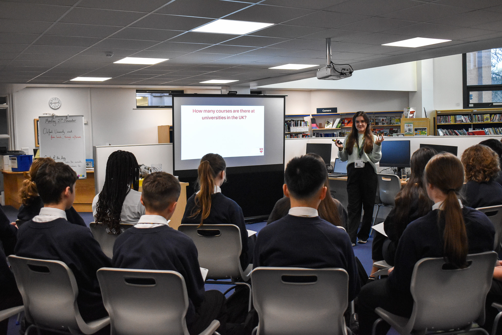 Molly speaking to pupils at Dowdales School. Image shows seating students from behind looking towards Molly standing at the front of the classroom next to a screen showing a powerpoint presentation asking how many university courses there are.