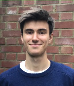 Matthew Buckley in a blue jumper with a brick wall background