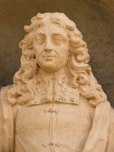 A close up of the head and shoulders of a sandy-coloured stone statue of Sir Joseph Williamson. His hair is shoulder-length and wavy, he wears a decorative square collar with lace detailing carved in, and decorative buttons can be seen on the front of his coat.