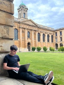 Josu Aurrekoetxea sitting in Front Quad with his laptop on his lap, typing. The clock tower and quad are in the background.