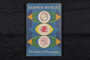 The book 'The Doors of Perception' lying flat so the cover is face-up. The cover has a blue background, with three large abstract eye designs taking up most of the space. They have geometric designs within the pupils and have red, yellow, green, and orange details so each eye is different. The author and title are at the head and foot of the cover respectively, printed in white.