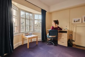 A student bedroom with a purple carpet, large bay window and desk to the right. A student sits at her laptop looking out the window. Sunlight pours through the window which has a small coffee table with books piled up.