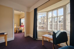 A student bedroom with a purple carpet, large bay window and desk to the right. A student sits in the ante room with a book and looks out the window.