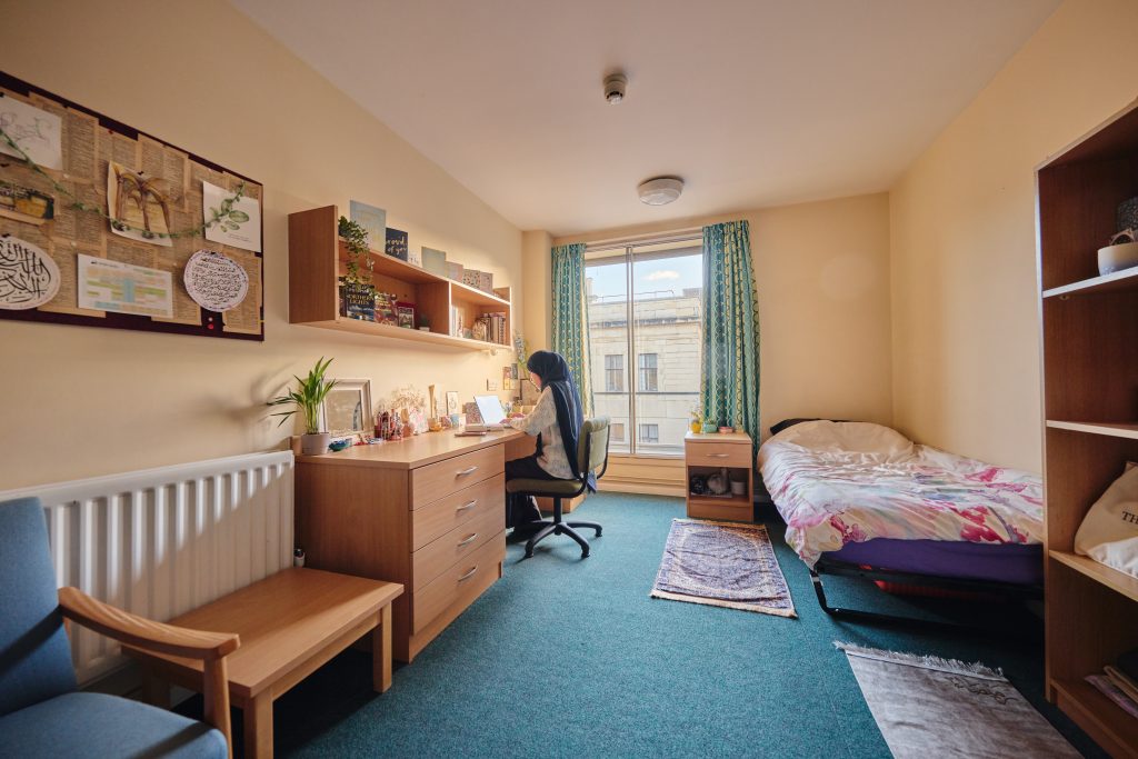 Student bedroom Student bedroom at St Aldate's House. There's a bed, tall bookshelf, chest of drawers, comfy chair, low table, desk and chair. A student sits at the desk typing. There's a large window at the end of the room.