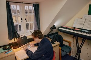 A student sits at his desk working on a laptop. There is a keyboard next to him and a window showing glimpses of Front Quad in front of him.