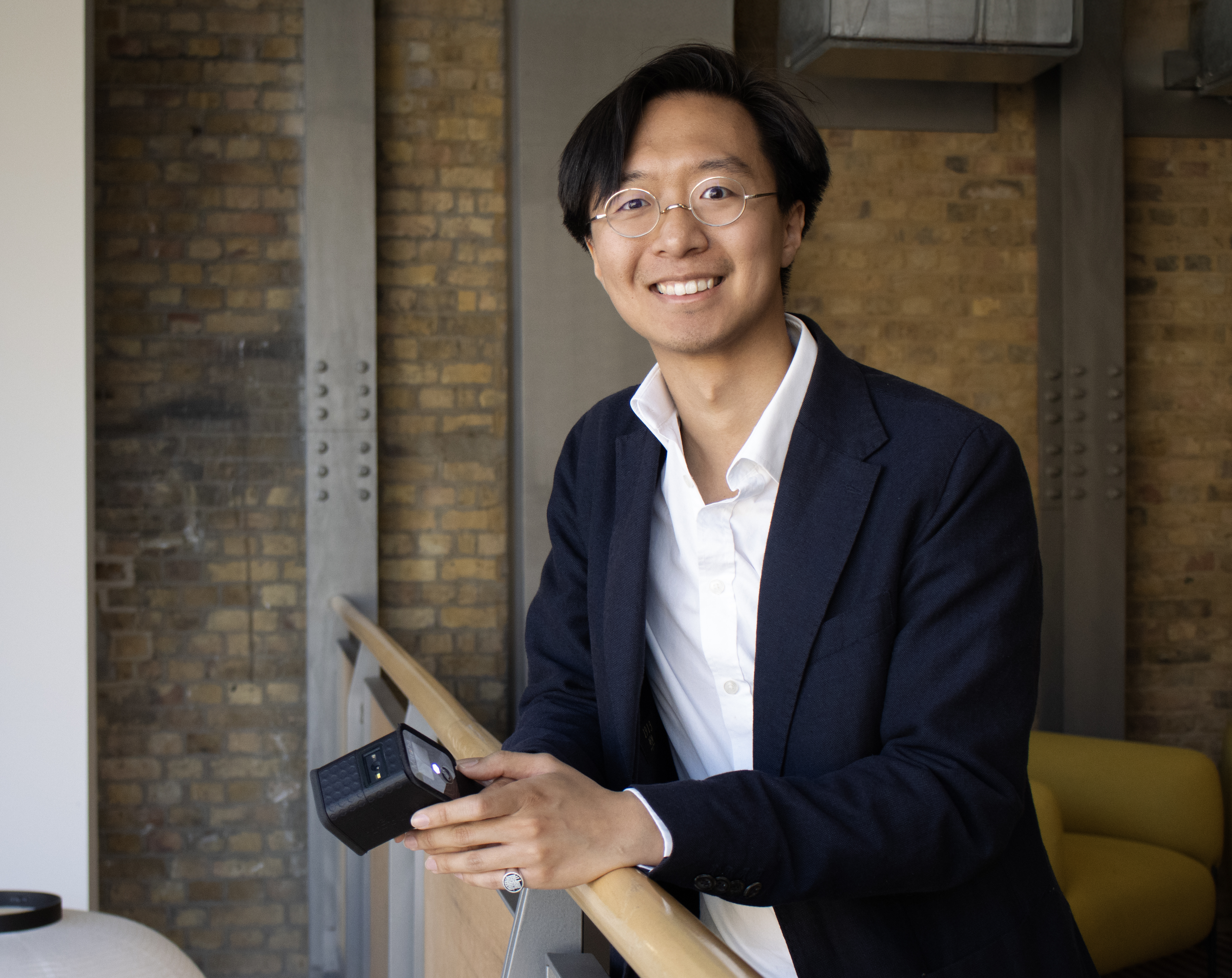 Hans Chan profile photo. Hans wears a dark suit jacket and a white shirt. He leans on a bannister and smiles to camera.