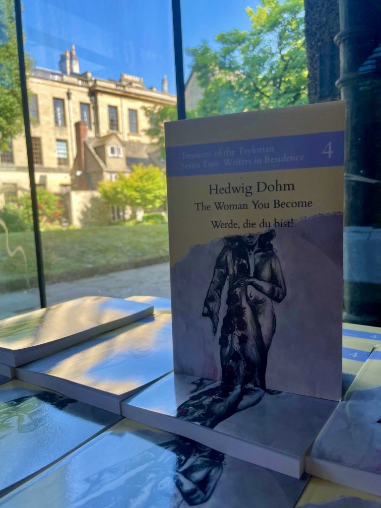 copies of the book 'The Woman You Become' piled up on a table in the foyer of the Shulman Auditorium. The Drawda garden is visible through the window.
