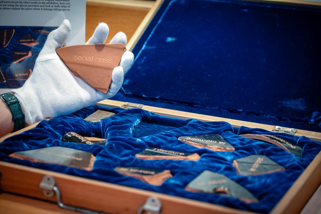 an open case with a blue velvet interior and inside the case sit fragments of pottery with words on them. One fragment has been lifted out and held up by a white-gloved hand and the words on it read 'social class'