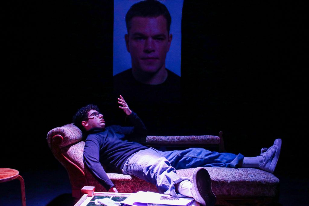 Production photos from 'Unprofessional' photo from the production of Unprofessional: a man is lying on a chaise langue looking at his hand, there's a photo of Matt Damon on the wall behind him