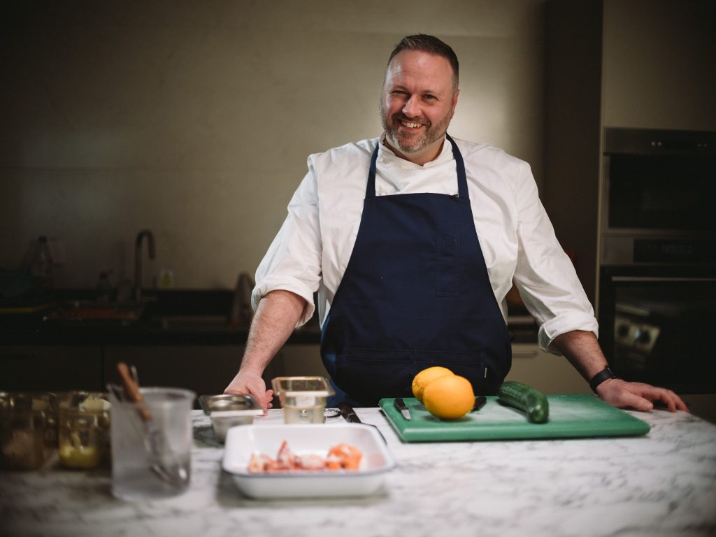 Sean Ducie stands with his hands resting on a counter top with a chopping board, food and utensils in front of him.