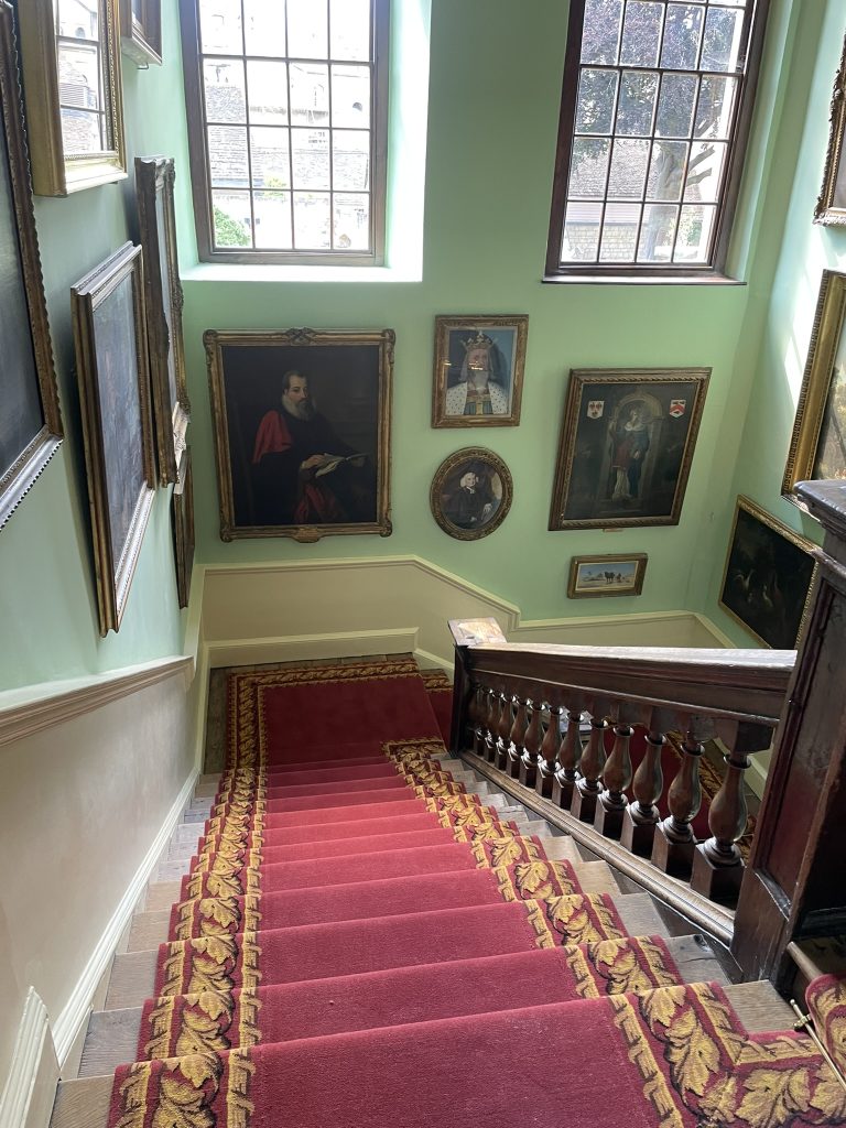The new SCR carpet: photo shows a staircase looking from above with a red carpet with golf leaf-patterned border. The walls are Cambridge blue and covered with portraits.