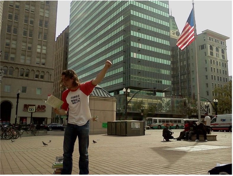 Poet David Brazil reading from a book with his arm raised outside in a US city