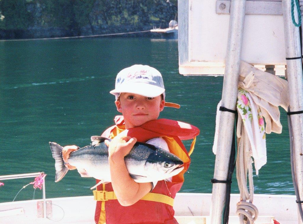 A photo of Peter fishing as a child. He is standing on a boat wearing a life jacket and holding a large fish