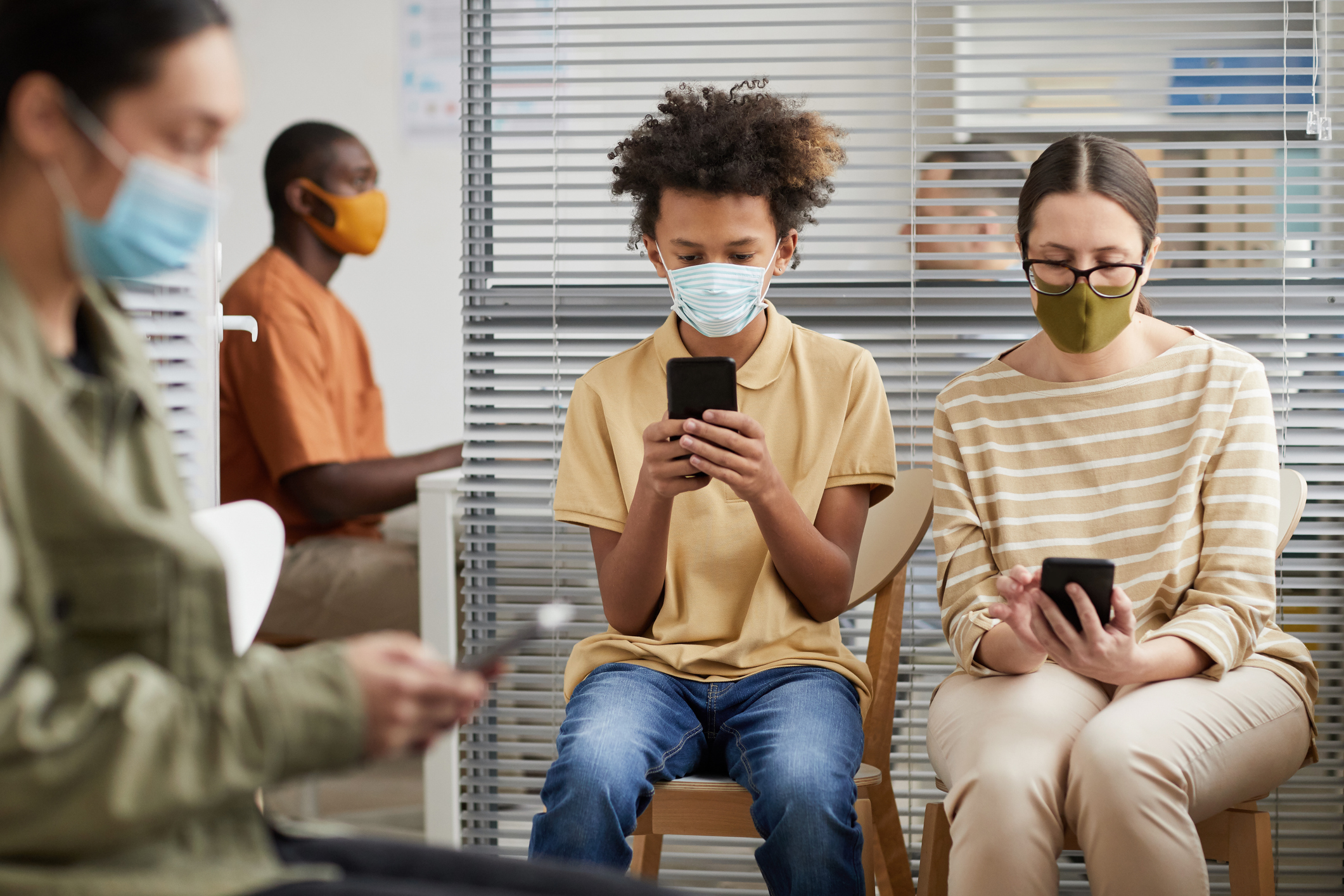 Front view portrait of multi-ethnic group of people using smartphones while waiting in line at medical clinic, all wearing masks. Credit: SeventyFour