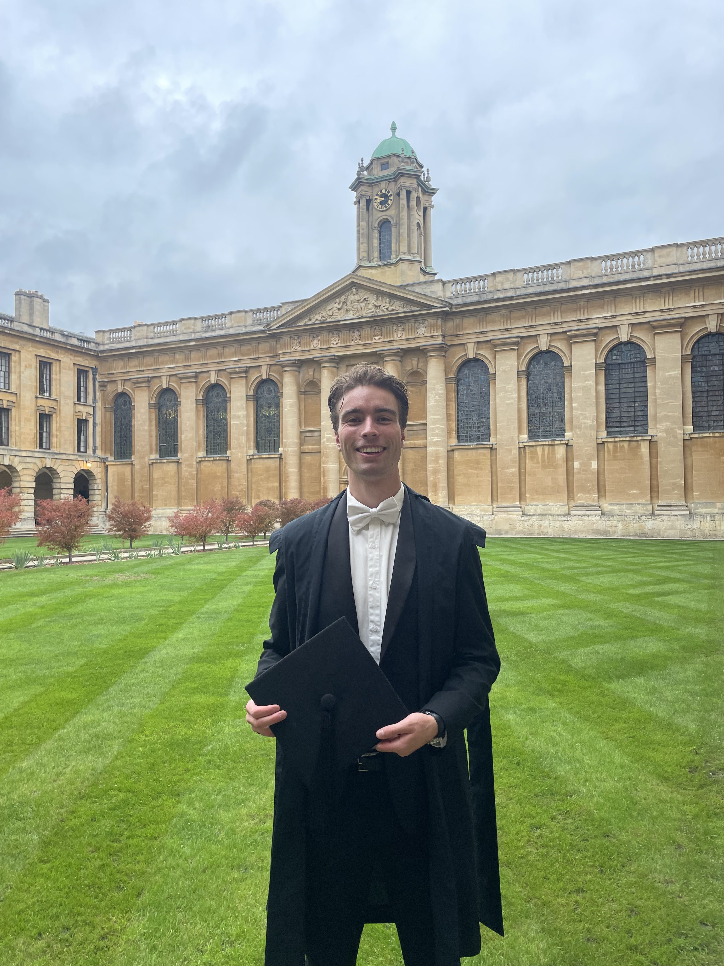 Billy pictured wearing academic dress and holding a mortarboard in the Front Quad