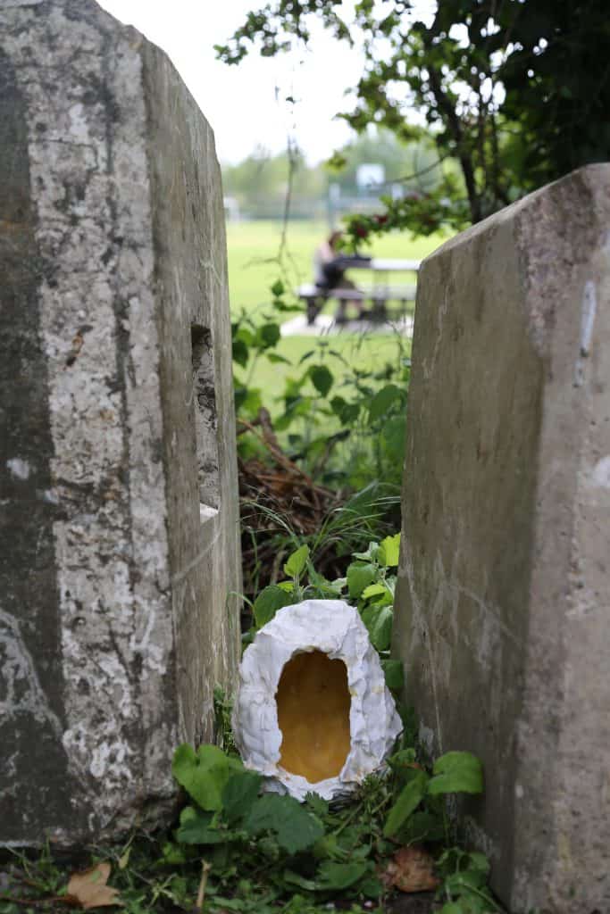 'Talking Head'​ (Plaster cast in clay, wax) by Maia Szulta​; image depicts a small plaster egg-shaped object with a hollowed out centre wedged between two stones, potentially gravestones with a field and picnic bench in the background