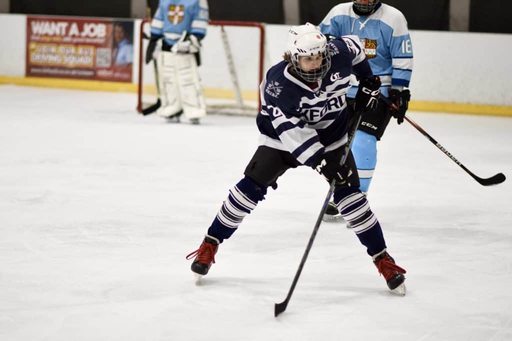 ice hockey player Aidan in a dark blue kit skates forward with the puck during a Varsity match, with two Cambridge players visible in the background.