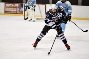 ice hockey player Aidan in a dark blue kit skates forward with the puck during a Varsity match, with two Cambridge players visible in the background.