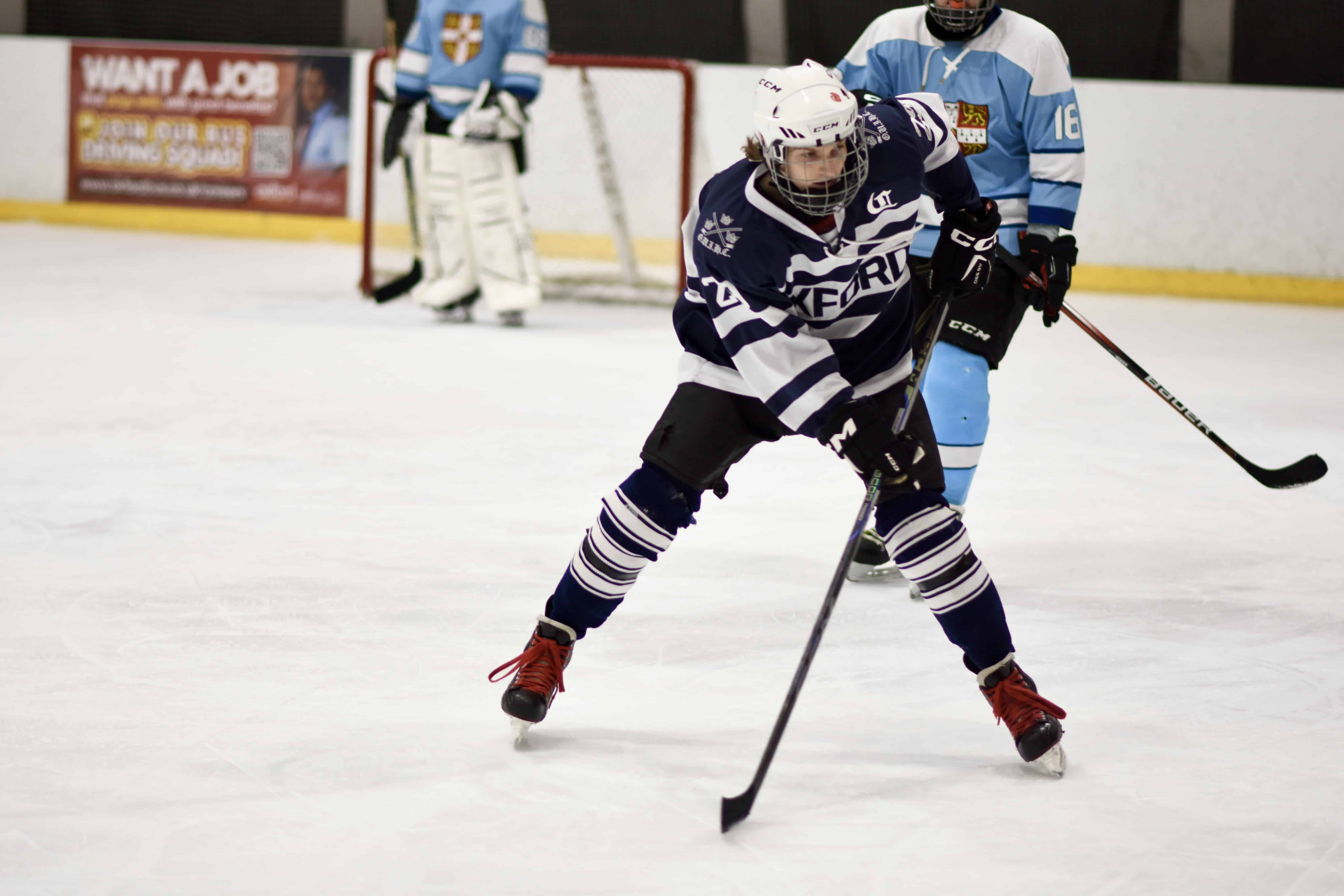 ice hockey player Aidan in a dark blue kit skates forward with the puck during a Varsity match, with two Cambridge players visible in the background.