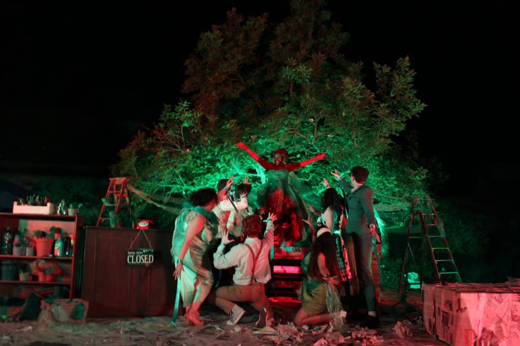 a production shot from an open air play in the gardens of Queen's College. The image is taken after dark and people are crowded around a green light
