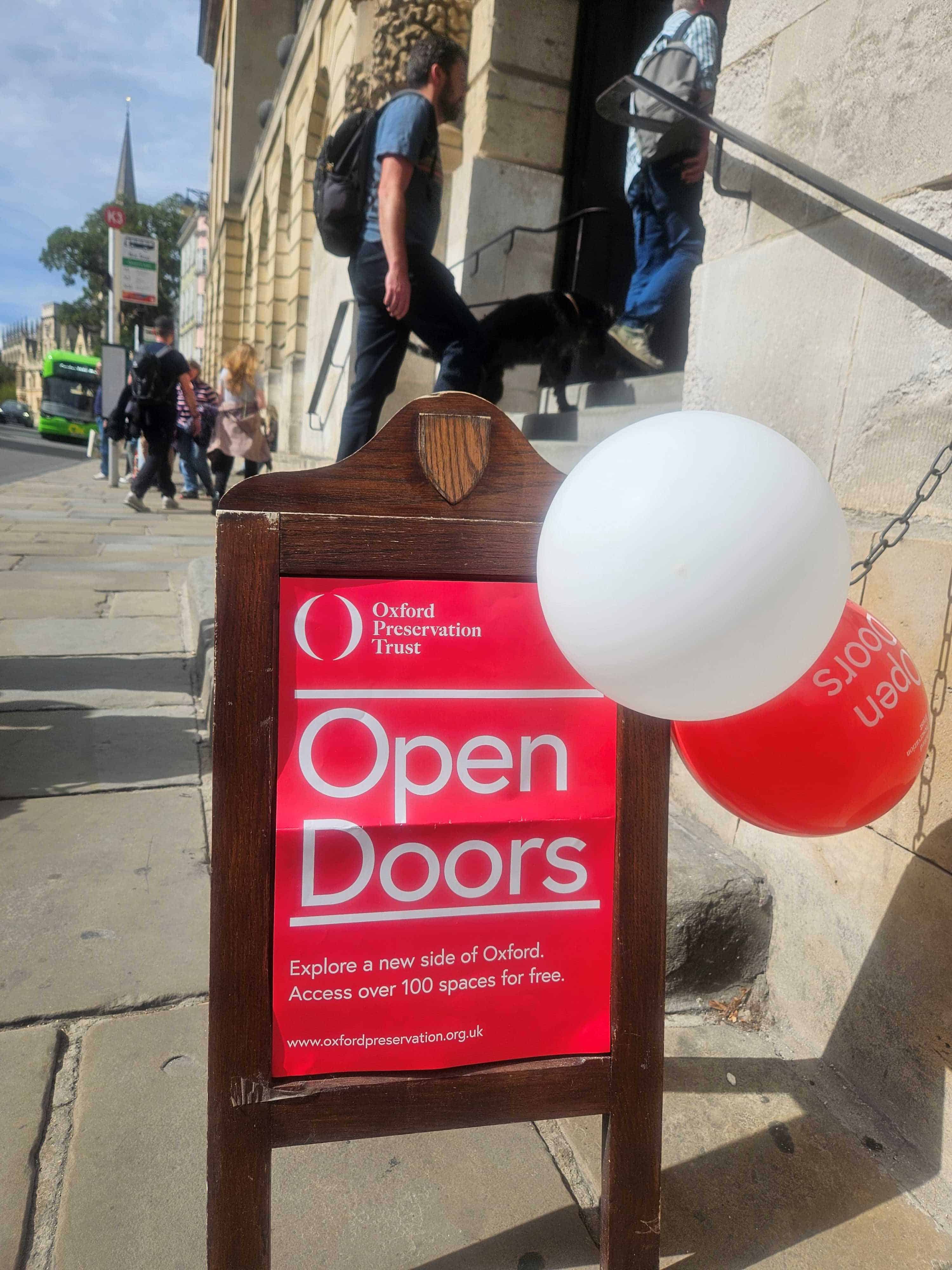 A red Oxford Open Doors poster on an a-frame at the foot of the Queen's College stairs on the High Street. Someone is walking up the steps and there's a red balloon and a white balloon attached to the a-frame.