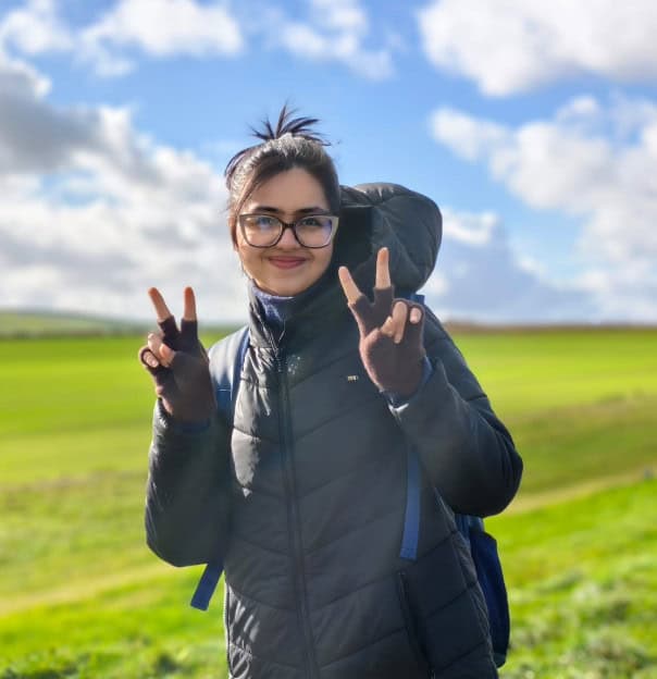 A photo of Jennice in a green field against a blue sky. She is giving the peace sign with both hands.