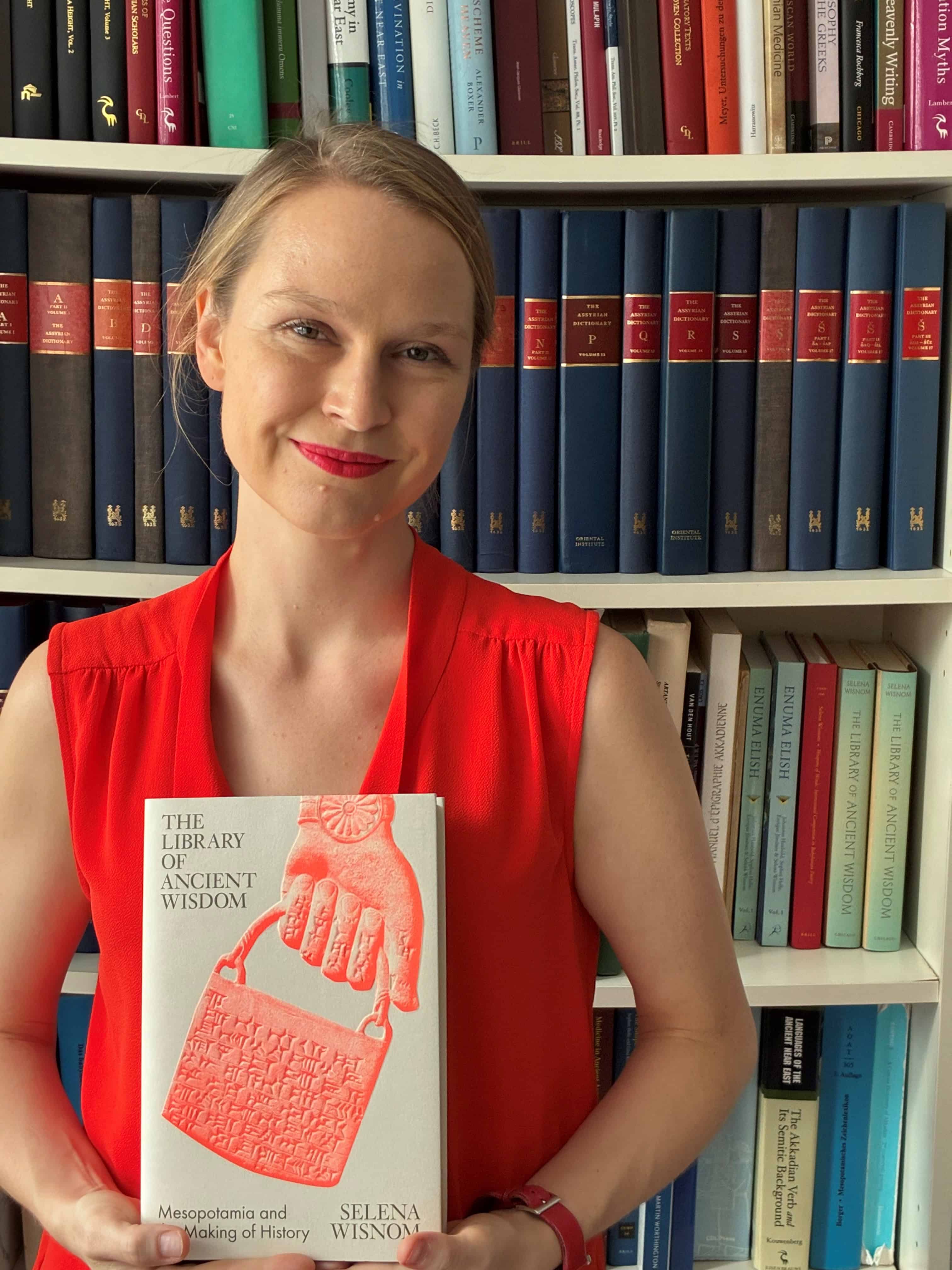 Dr Selena Wisnom in front of a bookcase holding her book 'The Library of Ancient Wisdom', the cover of which shows a stone tablet in handbag form being held by a stone-carved hand